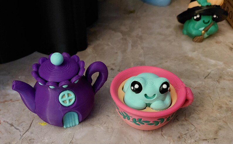 May include: A purple teapot with a light blue door and a matching lid, next to a pink teacup with a light blue frog figurine. A green frog wearing a witch hat is in the background. Whimsical, decorative items.