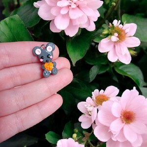 Puede incluir: Un pequeño amuleto de ratón de arcilla polimérica gris que sostiene un pequeño corazón naranja. El amuleto se sostiene en una mano sobre un fondo de flores rosas.