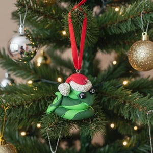 May include: A festive Christmas tree decoration featuring a green snake wearing a red Santa hat. The ornament is suspended by a red ribbon, amidst other baubles and lights on a Christmas tree.