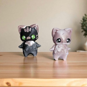 Może przedstawiać: Dwie figurki kotów, jedna czarna i jedna różowa, stoją obok siebie na drewnianej powierzchni. Czarny kot ma zielone oczy i białą pierś. Różowy kot ma duże, czarne oczy. Oba mają małe ramiona i kocie uszy.