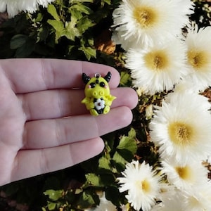 Op de afbeelding: Een handgemaakte gele drakenbedel met zwarte hoorns en vleugels, met een witte bloem met een zwart centrum. De bedel wordt in een hand gehouden, met witte bloemen op de achtergrond.