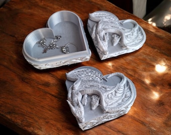 May include: Two heart-shaped trinket boxes with a dragon design. One box is open and shows a silver chain and ring inside.