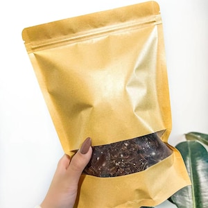 May include: A kraft paper stand-up pouch with a clear window, filled with dark brown contents. The pouch is held against a white background, with a glimpse of a green plant in the corner.