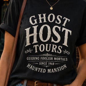 Może przedstawiać: Czarny t-shirt z białym napisem "GHOST HOST TOURS" i "HAUNTED MANSION". Koszulka ma podwinięte rękawy i jest połączona z niebieskimi dżinsami i brązowym paskiem. Tło zawiera znak z napisem "Haunted Mansion".