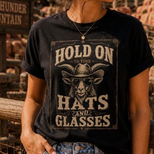 May include: A black short-sleeved t-shirt featuring a graphic print of a goat wearing a cowboy hat and sunglasses. The text "HOLD ON TO YOUR HATS AND GLASSES" is printed in a distressed font above and below the goat illustration on the front.