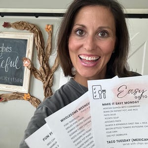 May include: A woman smiling and holding three printed menu sheets. The menus feature meal plans for the week, including options like tacos, pasta salad, and chicken dishes. The background includes a chalkboard with the words "ghes" and "eful".