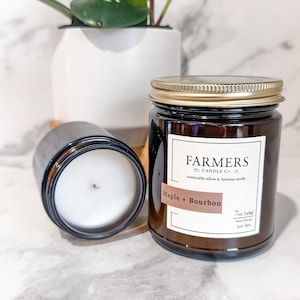 May include: A brown glass candle jar with a gold lid. The label reads "FARMERS CANDLE Co. LTD" and "sustainable tallow & beeswax candle". The candle scent is "Maple + Bourbon". The jar is 7 oz and has a burn time of 50+ hours.