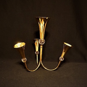 Puede incluir: Un candelabro de metal dorado con tres brazos y cuatro portavelas en forma de trompeta. Cada portavelas tiene un adorno de cristal transparente en la base.