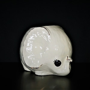 May include: White ceramic elephant planter with black eyes and a gold rim. The planter is shaped like an elephant's head and has a large opening at the top for planting.