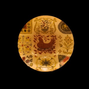 May include: A ceramic plate with a cream background and a colourful floral design. The design features a stylised bird in the centre, surrounded by various floral motifs and geometric patterns in shades of green, brown, red, and yellow.