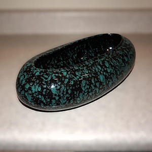 May include: An oval-shaped, speckled ceramic bowl. The bowl is black with teal-colored speckles throughout. The interior of the bowl is also black with teal speckles. The bowl has a glossy finish and is likely used for decorative purposes.