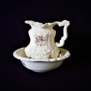 May include: A white ceramic pitcher and bowl set with floral designs. The pitcher has a ruffled rim and a handle. The bowl has a scalloped edge.