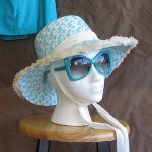 May include: A white and turquoise patterned sun hat with a white ribbon and fringe trim. The hat is worn on a white mannequin head with turquoise sunglasses.