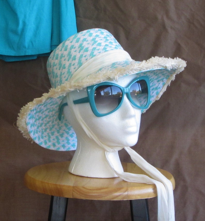 May include: A white and turquoise patterned sun hat with a white ribbon and fringe trim. The hat is worn on a white mannequin head with turquoise sunglasses.