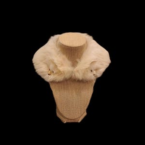 May include: Cream-colored fur collar displayed on a mannequin. The collar is fluffy and appears to be made of soft fur. The mannequin is a neutral tan color, and the background is black.