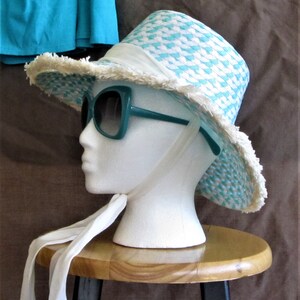 May include: A white and turquoise straw hat with a frayed brim and a white ribbon. The hat is worn by a white mannequin with teal sunglasses. The mannequin is sitting on a wooden stool.