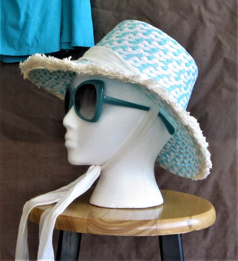 May include: A white and turquoise straw hat with a frayed brim and a white ribbon. The hat is worn by a white mannequin with teal sunglasses. The mannequin is sitting on a wooden stool.
