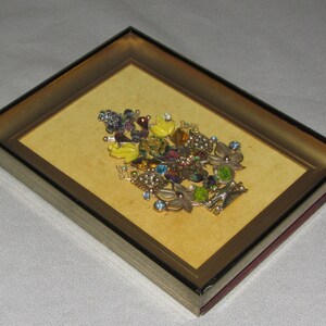 May include: A framed artwork featuring a colorful floral arrangement made of various rhinestones and jewels. The piece is set against a yellow background and framed in a gold-toned frame.