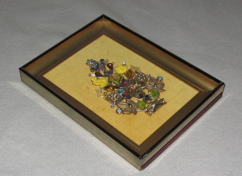 May include: A framed artwork featuring a colorful floral arrangement made of various rhinestones and jewels. The piece is set against a yellow background and framed in a gold-toned frame.