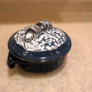 May include: A blue ceramic casserole dish with a white floral design and a unique, twisted handle. The dish has a glossy finish and is perfect for serving a variety of dishes.