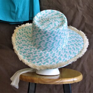 May include: A white and turquoise straw hat with a frayed brim. The hat is sitting on a wooden stool.