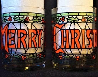 May include: Two clear glass tumblers featuring a stained glass style design with black outlines. The left glass displays the word MERRY in orange, and the right glass displays CHRIST in orange. Both feature green holly leaves and red berries.