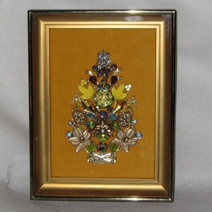 May include: A framed artwork featuring a colorful floral design made from various rhinestones and jewels. The design is set against a gold background and framed in a gold frame.