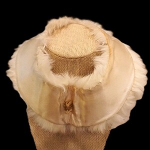 May include: Cream-colored vintage hat with a rounded crown and a wide brim. The hat is trimmed with soft, fluffy feathers and a satin ribbon. The hat is displayed on a tan mannequin head against a black background.