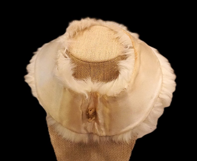 May include: Cream-colored vintage hat with a rounded crown and a wide brim. The hat is trimmed with soft, fluffy feathers and a satin ribbon. The hat is displayed on a tan mannequin head against a black background.