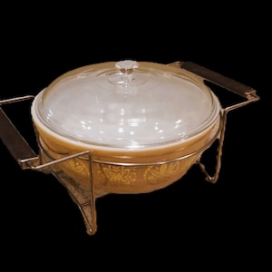 May include: A vintage brown casserole dish with a gold floral pattern and a glass lid. The dish is sitting on a metal stand with handles.
