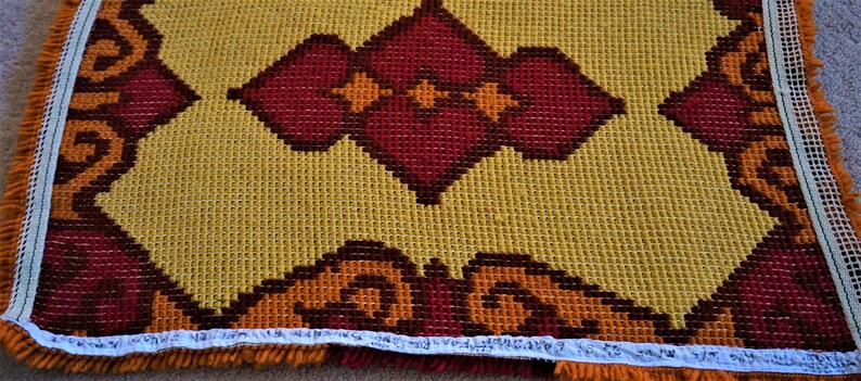 May include: A close-up of a yellow, orange, and red rug with a geometric pattern. The rug has a fringe border.