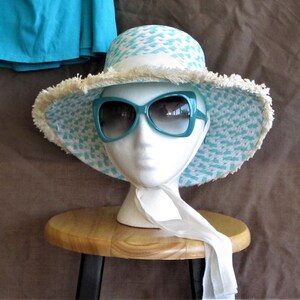 May include: A turquoise and white patterned sun hat with a white fringe trim and a white scarf tied around the neck of a white mannequin head. The hat is sitting on a wooden stool.