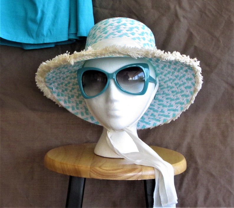 May include: A turquoise and white patterned sun hat with a white fringe trim and a white scarf tied around the neck of a white mannequin head. The hat is sitting on a wooden stool.