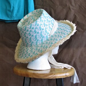 May include: A white and turquoise straw hat with a white ribbon and a fringe trim. The hat is sitting on a wooden stool.