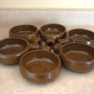 May include: Six brown ceramic bowls with handles are arranged on a light-colored surface. The bowls have a rounded shape with a small handle extending from the side. The bowls appear to be handmade and have a rustic aesthetic.