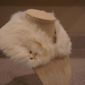 May include: Cream-colored faux fur collar with a small, decorative animal face. The collar is displayed on a mannequin head, showcasing its shape and texture. The fur appears soft and fluffy, adding a touch of elegance.