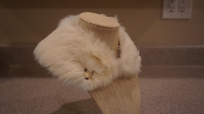May include: Cream-colored faux fur collar with a small, decorative animal face. The collar is displayed on a mannequin head, showcasing its shape and texture. The fur appears soft and fluffy, adding a touch of elegance.