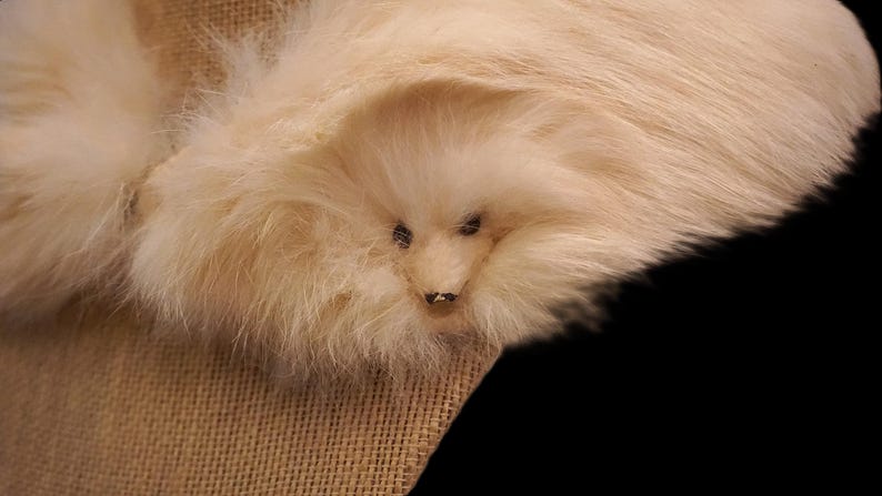 May include: A close-up of a plush, cream-colored animal head with small, dark eyes and a tiny nose. The fur is soft and fluffy, with a slightly darker shade around the eyes. The animal is resting on a tan fabric.