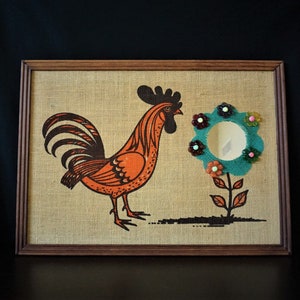 May include: A framed artwork featuring a rooster with an orange and black design, standing next to a blue flower with a mirror in the center. The flower has colorful accents around the mirror.