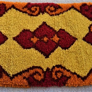 May include: A small, rectangular rug with a geometric pattern in shades of orange, yellow, and red. The rug has a fluffy texture and is made of yarn.