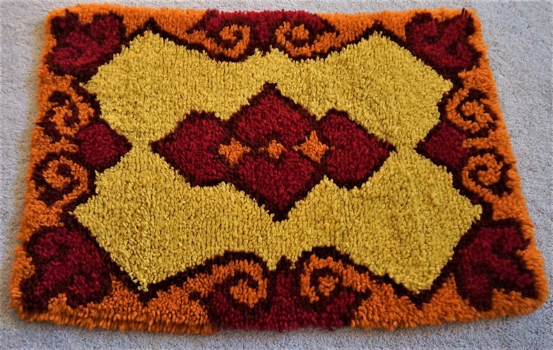 May include: A small, rectangular rug with a geometric pattern in shades of orange, yellow, and red. The rug has a fluffy texture and is made of yarn.