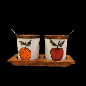 May include: A set of two white ceramic jars with wooden lids and small spoons, resting on a wooden tray. Each jar features a hand-painted fruit design: an orange and a red apple. The background is black.