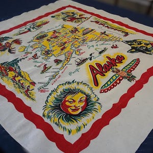 Puede incluir: Un pañuelo de recuerdo vintage blanco y rojo de Alaska. El pañuelo presenta un mapa colorido de Alaska con ilustraciones de lugares emblemáticos locales, vida silvestre y la palabra "Alaska" en una fuente estilizada. El borde es rojo.