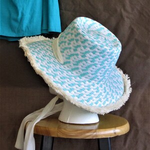 May include: A white and turquoise straw sun hat with a white trim and a white ribbon. The hat is sitting on a wooden stool.