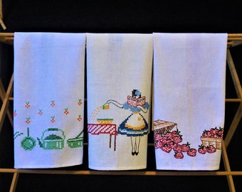 May include: Three white tea towels with embroidered designs. The first towel has a green tea pot and two cups. The second towel has a woman holding a cookbook and a table with a red and white checkered tablecloth. The third towel has a basket of red strawberries.