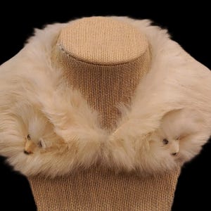 May include: Cream-colored faux fur collar with two animal-shaped accents. The collar is displayed on a tan mannequin head against a black background. The fur appears soft and fluffy, suggesting a vintage or retro style.