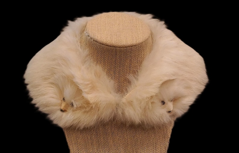 May include: Cream-colored faux fur collar with two animal-shaped accents. The collar is displayed on a tan mannequin head against a black background. The fur appears soft and fluffy, suggesting a vintage or retro style.