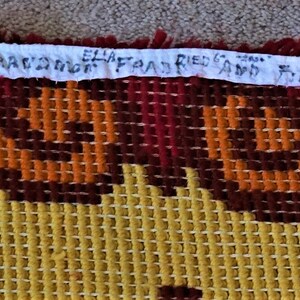 May include: A close-up of a colorful woven tapestry with a geometric pattern in shades of orange, brown, and yellow. The tapestry has a fringe border and a handwritten label that reads "MADE IN SPAIN 63 X 63" and "FRANKFURT 61 HELLEN BACON DOOR".