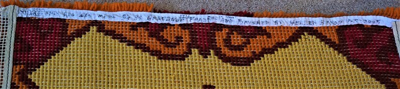 May include: A close-up of a colorful woven tapestry with a geometric pattern in shades of orange, brown, and yellow. The tapestry has a fringe border and a handwritten label that reads "MADE IN SPAIN 63 X 63" and "FRANKFURT 61 HELLEN BACON DOOR".