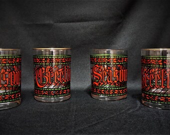 May include: Four clear glass tumblers featuring a stained glass pattern in red and green. The design includes a decorative border and the red text "Season's Greetings" wrapped around the exterior of each glass, with a gold rim at the top edge.